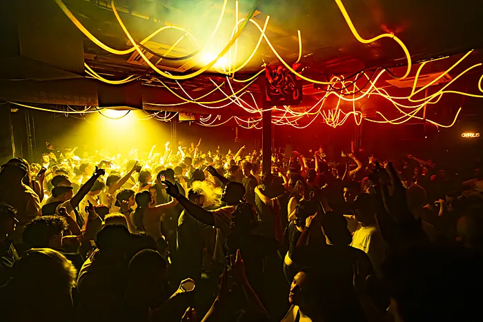 A packed XOYO basement under yellow light, hands up, neon tubes overhead.